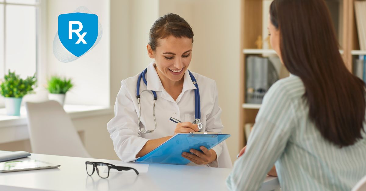 A female doctor in a white coat smiling at a female patient while writing on a clipboard in a bright medical office.