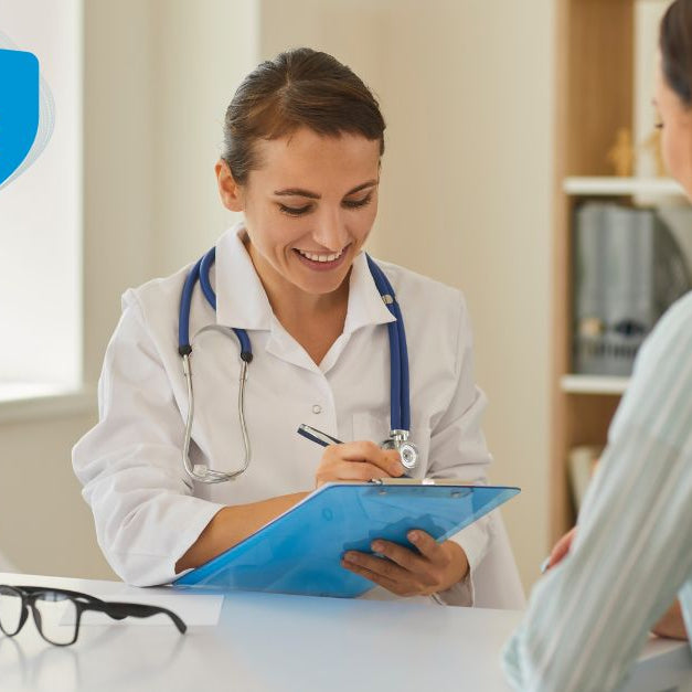 A female doctor in a white coat smiling at a female patient while writing on a clipboard in a bright medical office.