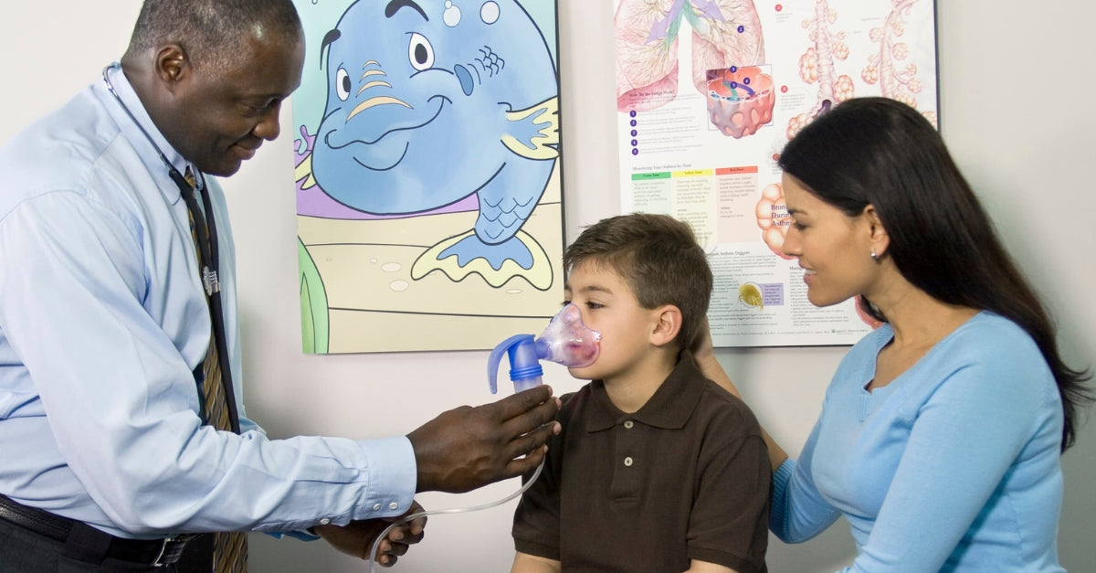 Alt text: "Doctor fitting a young boy with a nebulizer mask in an exam room, with his mother by his side.